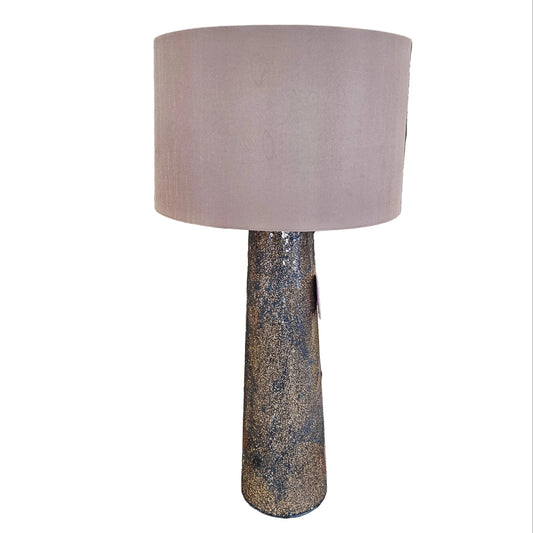 A table lamp with a large dusty pink fabric drum shade and a textured, speckled dark brown and gold base, shown in a cluttered furniture store setting.